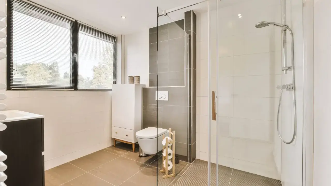 Salle de bain moderne avec une douche italienne, un WC suspendu et un meuble vasque. La lumière naturelle filtre à travers les stores.