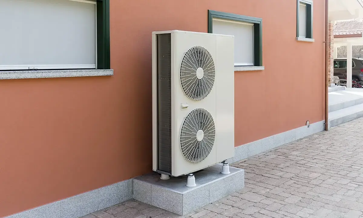 Unité extérieure de pompe à chaleur, avec ses deux ventilateurs, installée sur un socle devant un mur ocre. Le poids de cet appareil assure sa stabilité.