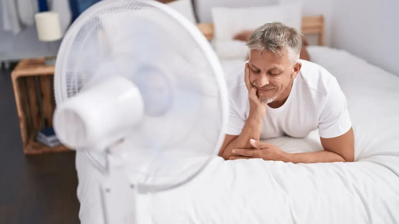Un homme se repose sur un lit blanc, profitant de la brise d'un ventilateur. Une astuce simple pour comment rafraîchir une chambre quand il fait chaud.