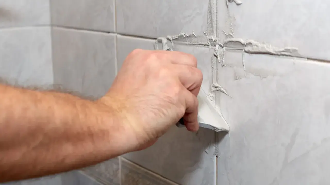 Main d'un bricoleur appliquant du mastic pour refaire joint carrelage sur un mur carrelé.