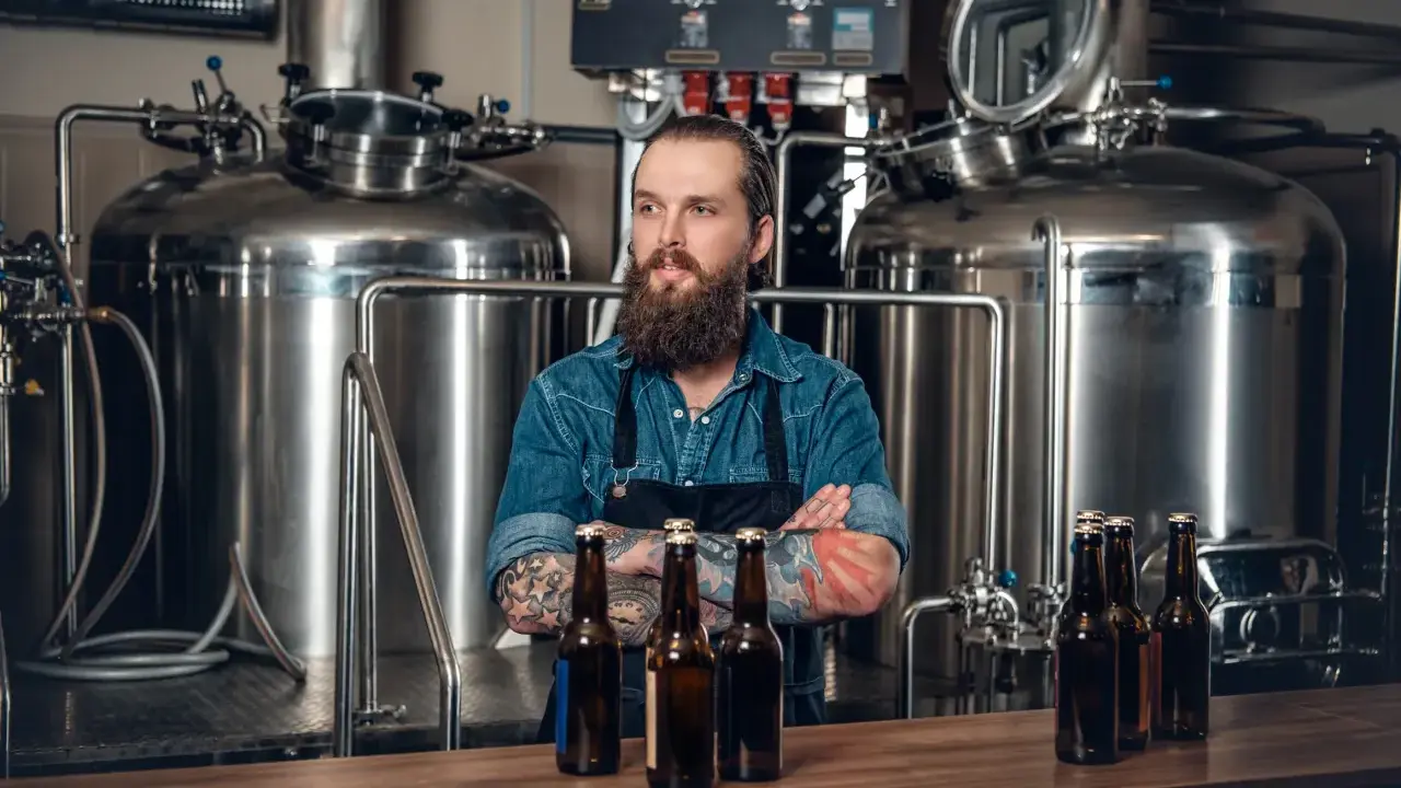 Un brasseur barbu pose devant une cuve chauffante, des bouteilles de bière sur le comptoir.