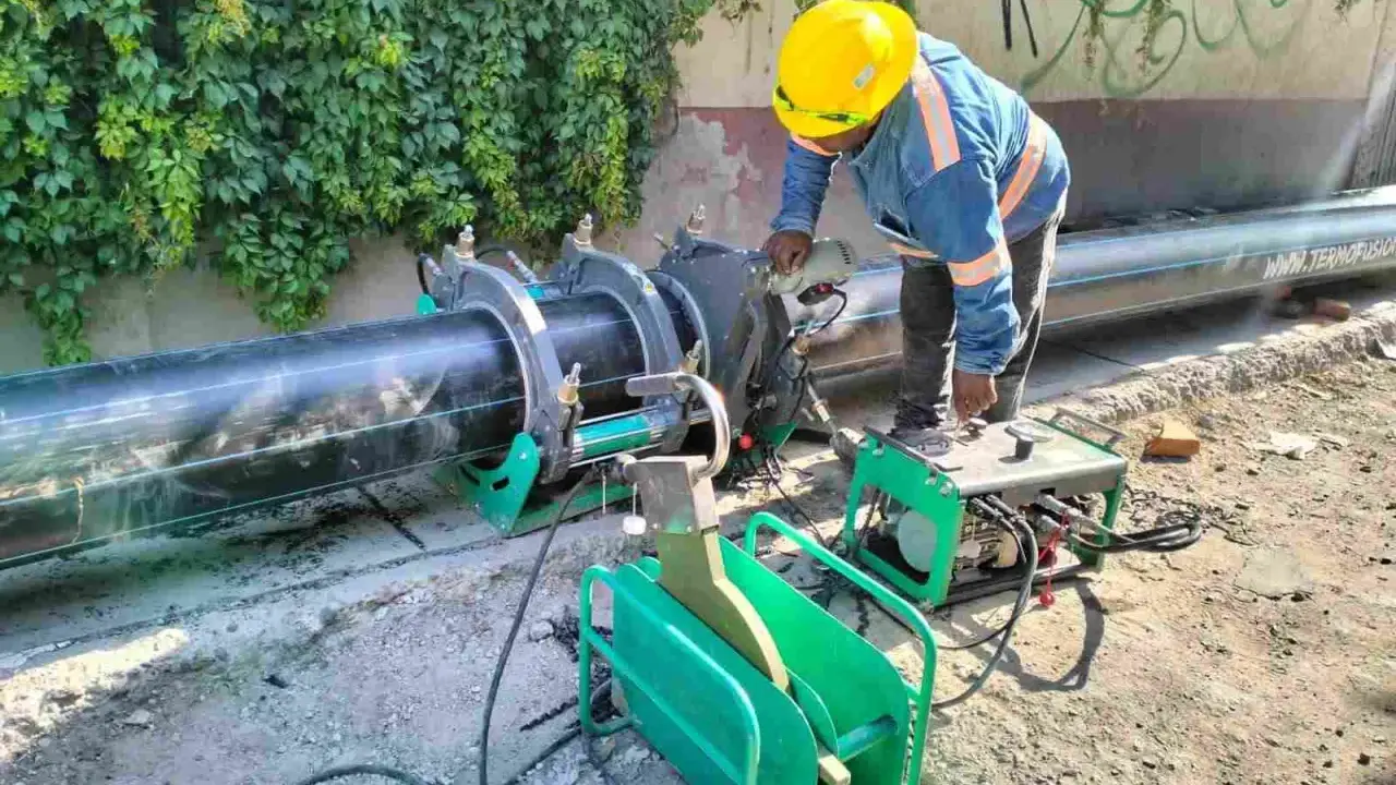 Un ouvrier utilise une machine de soudure pour joindre un gros tuyau noir. L'équipement comprend une douille à souder et une unité de contrôle verte.