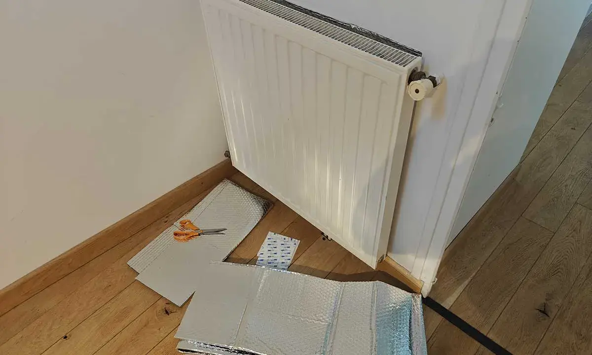 Un radiateur derrière une porte, prêt à être isolé avec des feuilles argentées et des ciseaux.