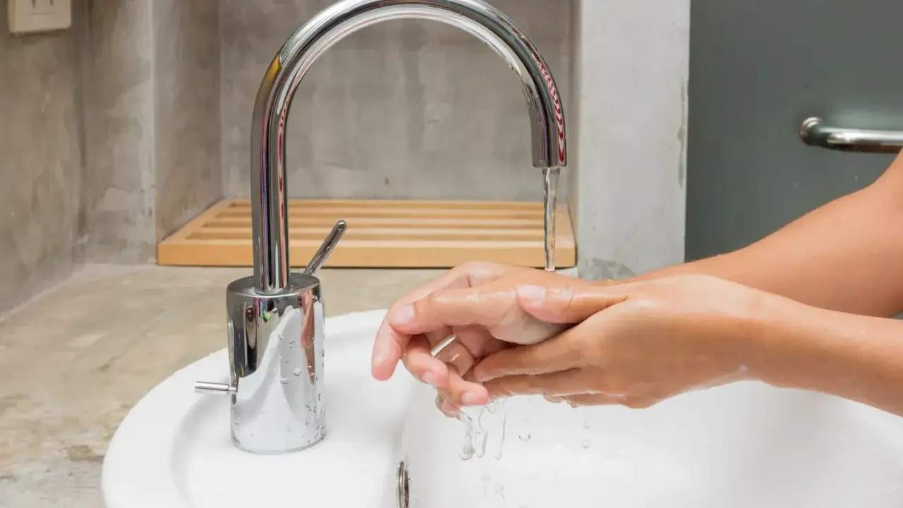Mains d'une personne se lavant sous un robinet moderne en inox. L'eau coule dans un lavabo blanc.