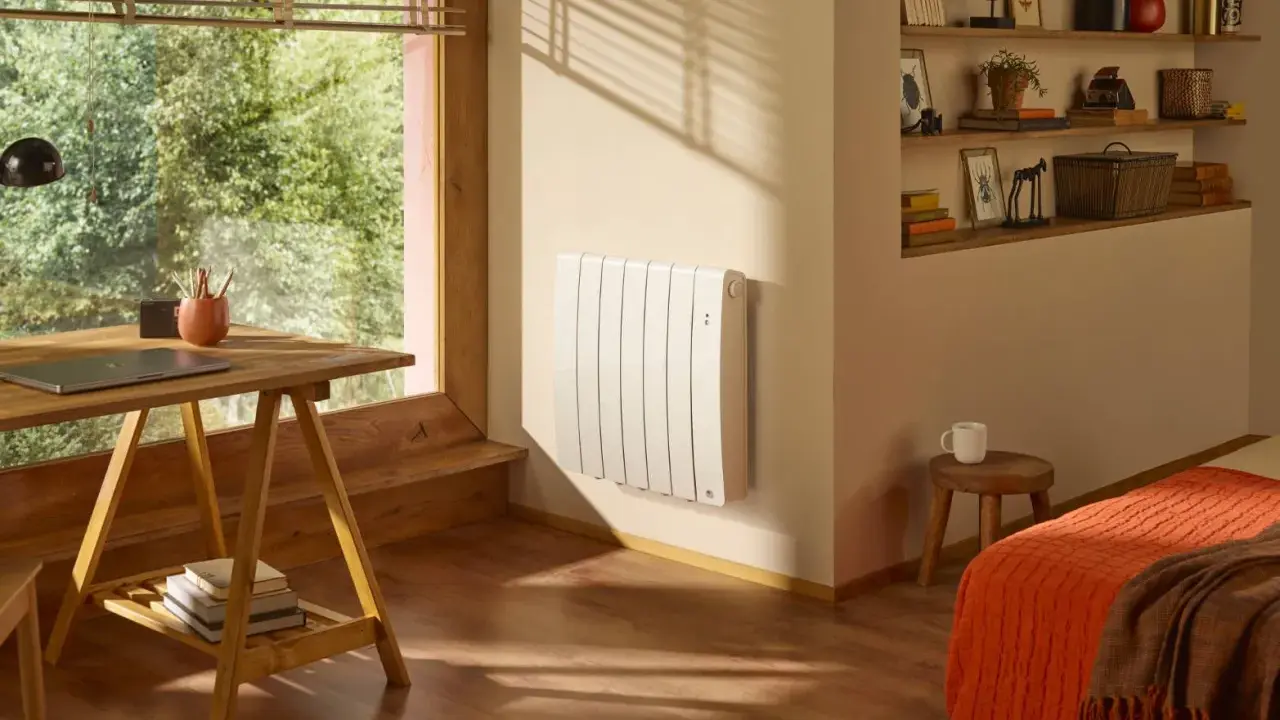 Un radiateur blanc vertical est fixé au mur, sous une fenêtre. Un bureau en bois et une étagère remplie d'objets complètent la pièce.