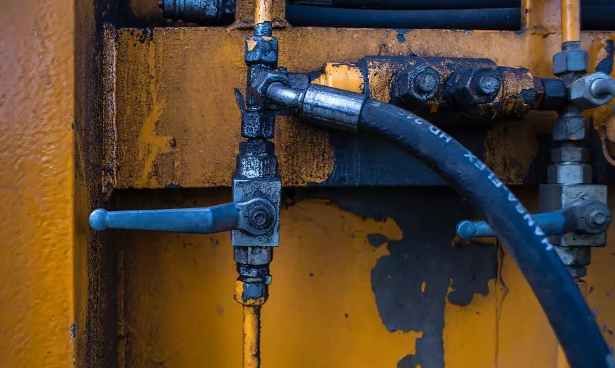 Détail d'un système hydraulique avec un tuyau noir marqué "HANSA-FLEX HD 24", un levier bleu et un sertissage flexible sur fond jaune usé.