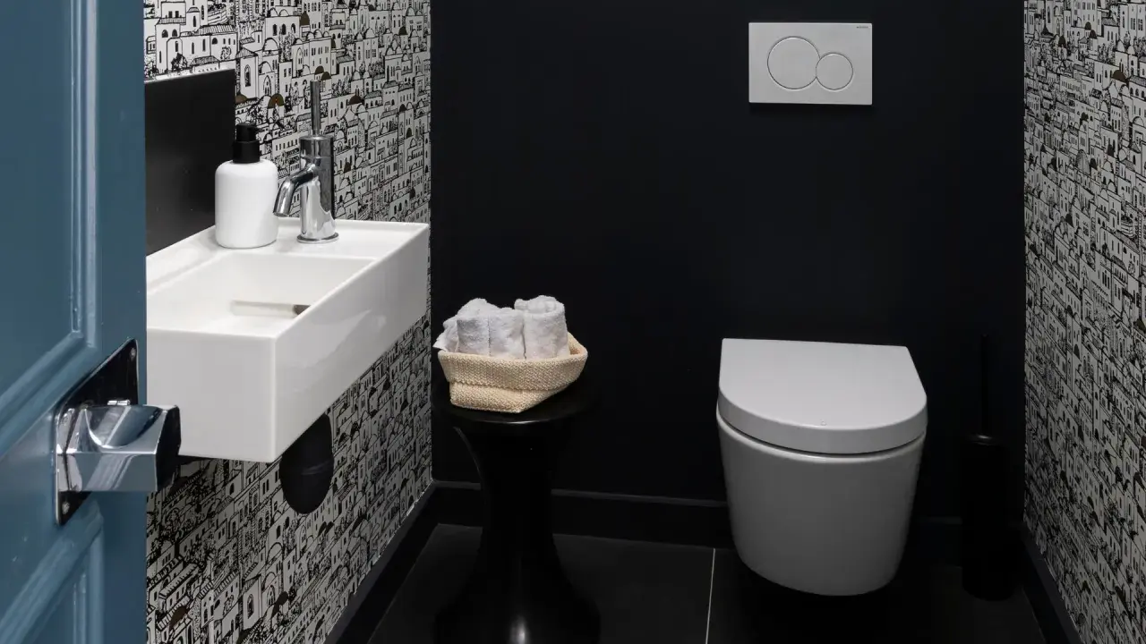 Une salle de bain moderne avec un papier peint original, un petit lavabo blanc, des serviettes roulées et une cuvette suspendue. La deco wc est soignée.