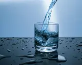 Un jet d'eau de source calcaire tombe dans un verre, créant des éclaboussures. Des gouttes d'eau parsèment la surface sombre.
