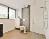 Salle de bain moderne avec une douche italienne, un WC suspendu et un meuble vasque. La lumière naturelle filtre à travers les stores.
