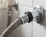 Gros plan sur un raccord double en métal chromé, connectant un tuyau tressé à une valve murale.