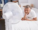 Un homme se repose sur un lit blanc, profitant de la brise d'un ventilateur. Une astuce simple pour comment rafraîchir une chambre quand il fait chaud.