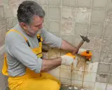Un homme en salopette jaune utilise un marteau pour enlever le carrelage mural, révélant le plâtre en dessous.