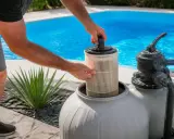 Entretien de la pompe à sable : une main retire le filtre d'une cuve grise, prêt pour le nettoyage. Piscine bleue en arrière-plan.