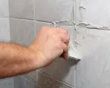 Main d'un bricoleur appliquant du mastic pour refaire joint carrelage sur un mur carrelé.