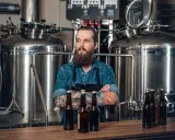 Un brasseur barbu pose devant une cuve chauffante, des bouteilles de bière sur le comptoir.