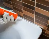 Application d'un joint silicone blanc à l'aide d'un pistolet, créant une barrière étanche entre le carrelage et une surface blanche.