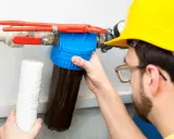 Un homme en casque jaune effectue la régénération manuelle d'un adoucisseur d'eau, tenant un nouveau filtre.
