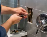 Plombier ajustant une pièce de robinetterie avec un tournevis. Un flexible renforcé est visible dans le mur.