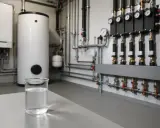 Un bécher d'eau claire devant un chauffe-eau et un système de tuyauterie complexe. Est-ce une eau dure ?