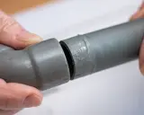 Mains de personne assemblant deux tuyaux gris en **pvc renforcé**, l'un avec un coude, prêts pour une installation solide.