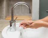 Mains d'une personne se lavant sous un robinet moderne en inox. L'eau coule dans un lavabo blanc.