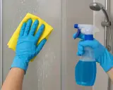 Mains gantées en bleu nettoient une paroi de douche entartrée avec un chiffon jaune. Un spray bleu est prêt à enlever le calcaire douche.