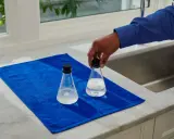 Main tenant une fiole pour mesurer th eau, posée sur une serviette bleue à côté d'une autre fiole.