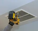 Main tenant utilise une visseuse pour l'entretien de la VMC, fixant la grille d'aération au plafond.