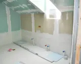 Travaux de finition dans une salle de bain : murs en plaques de plâtre, arrivées d'eau et niveau à bulle au sol. Isolation salle de bain en cours.