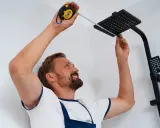 Un homme mesure avec un mètre ruban pour installer une colonne de douche noire dans une salle de bain carrelée.