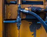 Détail d'un système hydraulique avec un tuyau noir marqué "HANSA-FLEX HD 24", un levier bleu et un sertissage flexible sur fond jaune usé.
