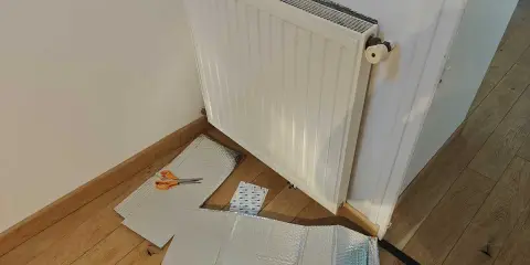 Un radiateur derrière une porte, prêt à être isolé avec des feuilles argentées et des ciseaux.