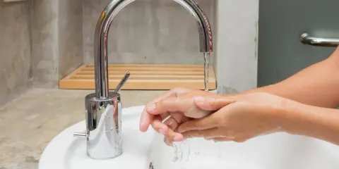 Mains d'une personne se lavant sous un robinet moderne en inox. L'eau coule dans un lavabo blanc.