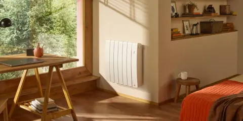 Un radiateur blanc vertical est fixé au mur, sous une fenêtre. Un bureau en bois et une étagère remplie d'objets complètent la pièce.