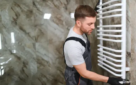 Un plombier effectue le branchement d'un sèche serviette blanc dans une salle de bain aux murs marbrés.