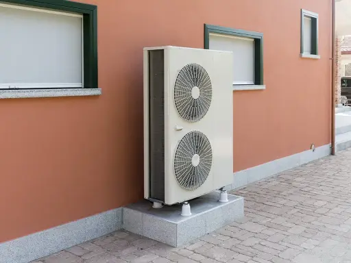 Unité extérieure de pompe à chaleur, avec ses deux ventilateurs, installée sur un socle devant un mur ocre. Le poids de cet appareil assure sa stabilité.