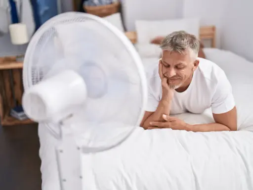 Un homme se repose sur un lit blanc, profitant de la brise d'un ventilateur. Une astuce simple pour comment rafraîchir une chambre quand il fait chaud.