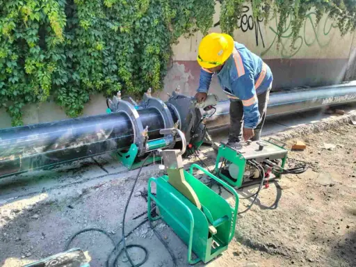 Un ouvrier utilise une machine de soudure pour joindre un gros tuyau noir. L'équipement comprend une douille à souder et une unité de contrôle verte.