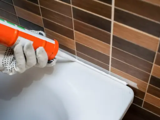 Application d'un joint silicone blanc à l'aide d'un pistolet, créant une barrière étanche entre le carrelage et une surface blanche.