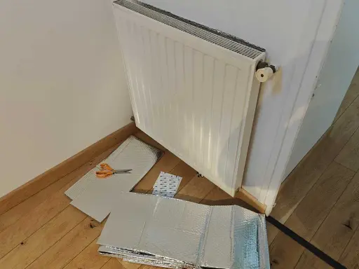 Un radiateur derrière une porte, prêt à être isolé avec des feuilles argentées et des ciseaux.