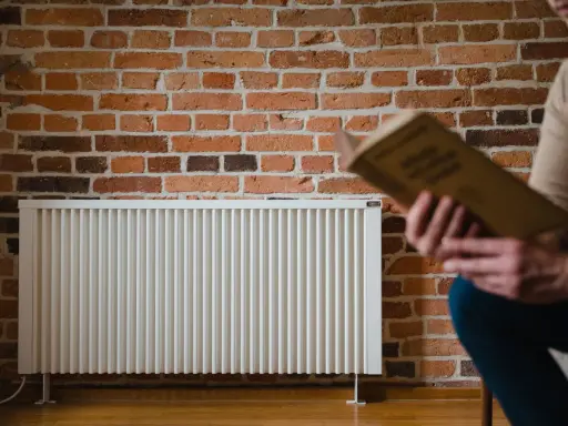 Un homme lit un livre près d'un radiateur blanc. La question quelle puissance de radiateur pour 25 m2 est au cœur de ce décor cosy.