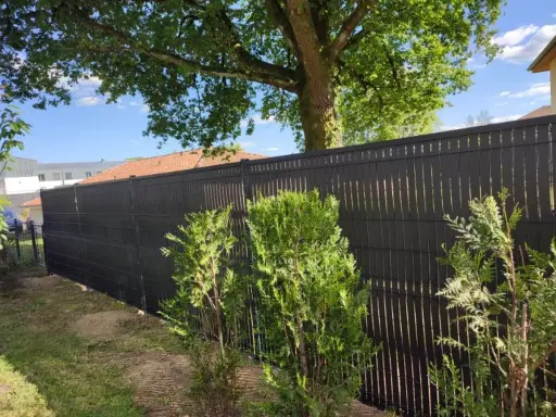 Clôture ajourée noire, offrant une protection discrète pour les tuyaux PVC. Des arbustes verts bordent cette barrière moderne.