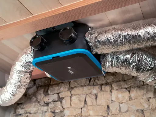 Unité de ventilation maison avec conduits argentés, installée sous un plafond en bois et au-dessus d'un mur en pierre.
