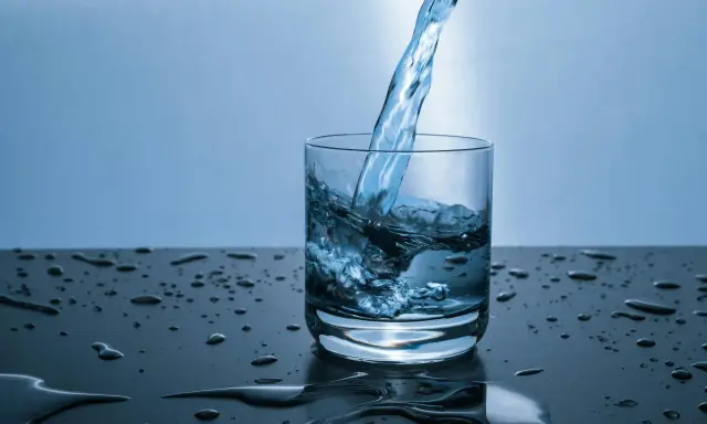 Un jet d'eau de source calcaire tombe dans un verre, créant des éclaboussures. Des gouttes d'eau parsèment la surface sombre.