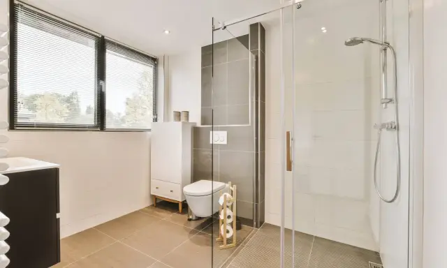 Salle de bain moderne avec une douche italienne, un WC suspendu et un meuble vasque. La lumière naturelle filtre à travers les stores.