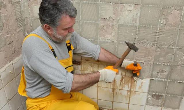 Un homme en salopette jaune utilise un marteau pour enlever le carrelage mural, révélant le plâtre en dessous.