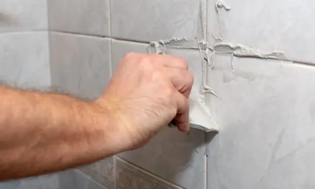 Main d'un bricoleur appliquant du mastic pour refaire joint carrelage sur un mur carrelé.