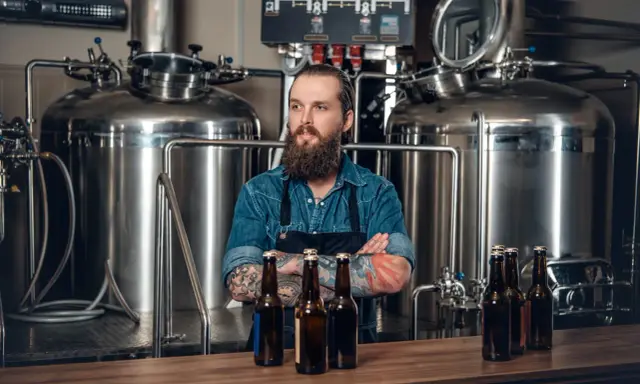 Un brasseur barbu pose devant une cuve chauffante, des bouteilles de bière sur le comptoir.