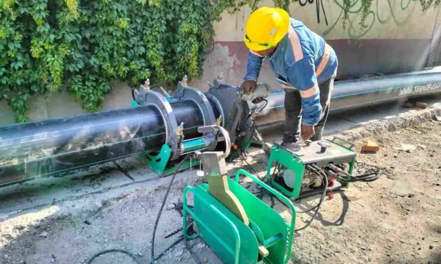 Un ouvrier utilise une machine de soudure pour joindre un gros tuyau noir. L'équipement comprend une douille à souder et une unité de contrôle verte.