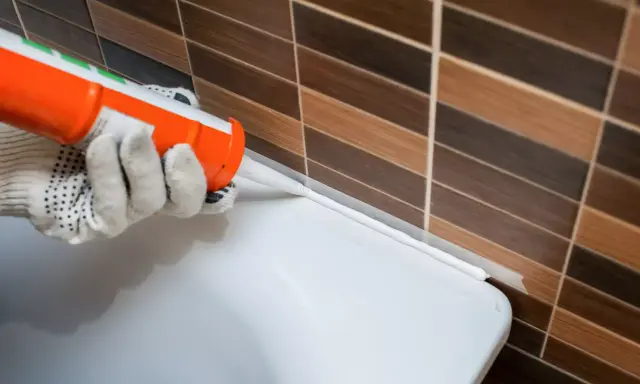 Application d'un joint silicone blanc à l'aide d'un pistolet, créant une barrière étanche entre le carrelage et une surface blanche.